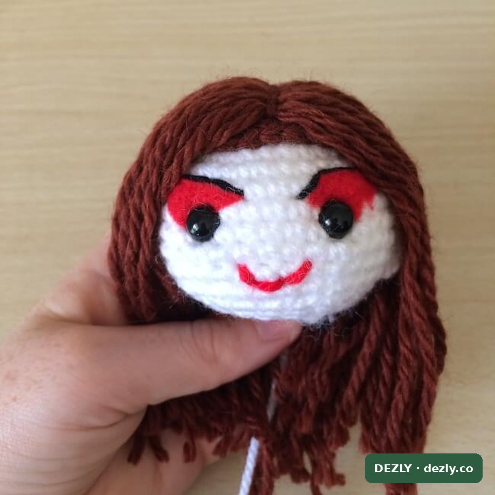 La cabeza terminada con la larga melena marrón óxido cayendo sobre los hombros, el maquillaje rojo y la sonrisa roja bordada.