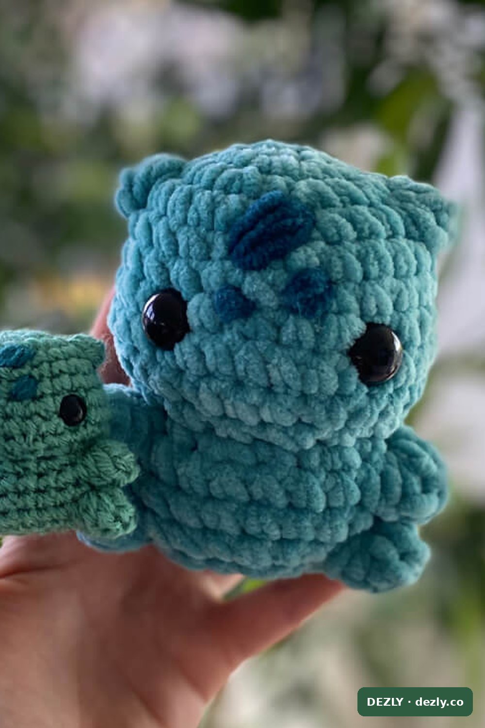 Two crocheted plush dolls held in one hand: a large one worked in teal chenille yarn beside a smaller one in smooth yarn, showing the plush size chenille creates.